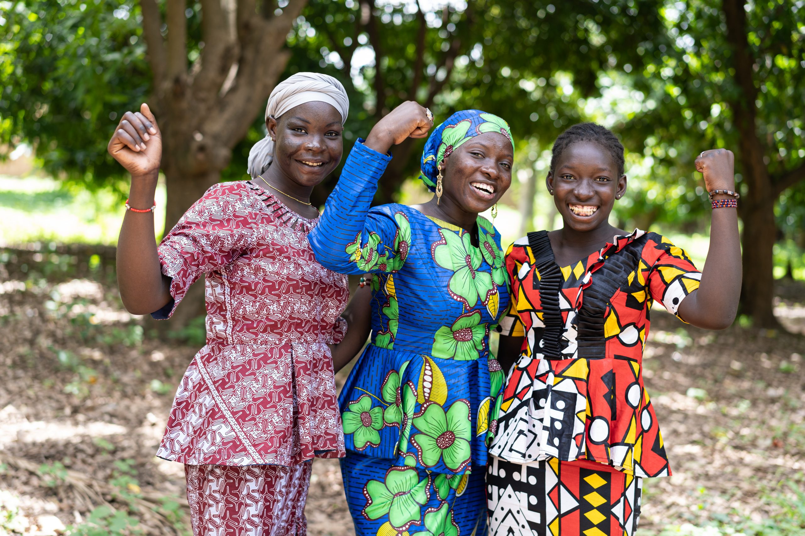 Three young black African villagers in colourful traditional dresses smiling at the camera with their clenched fists.