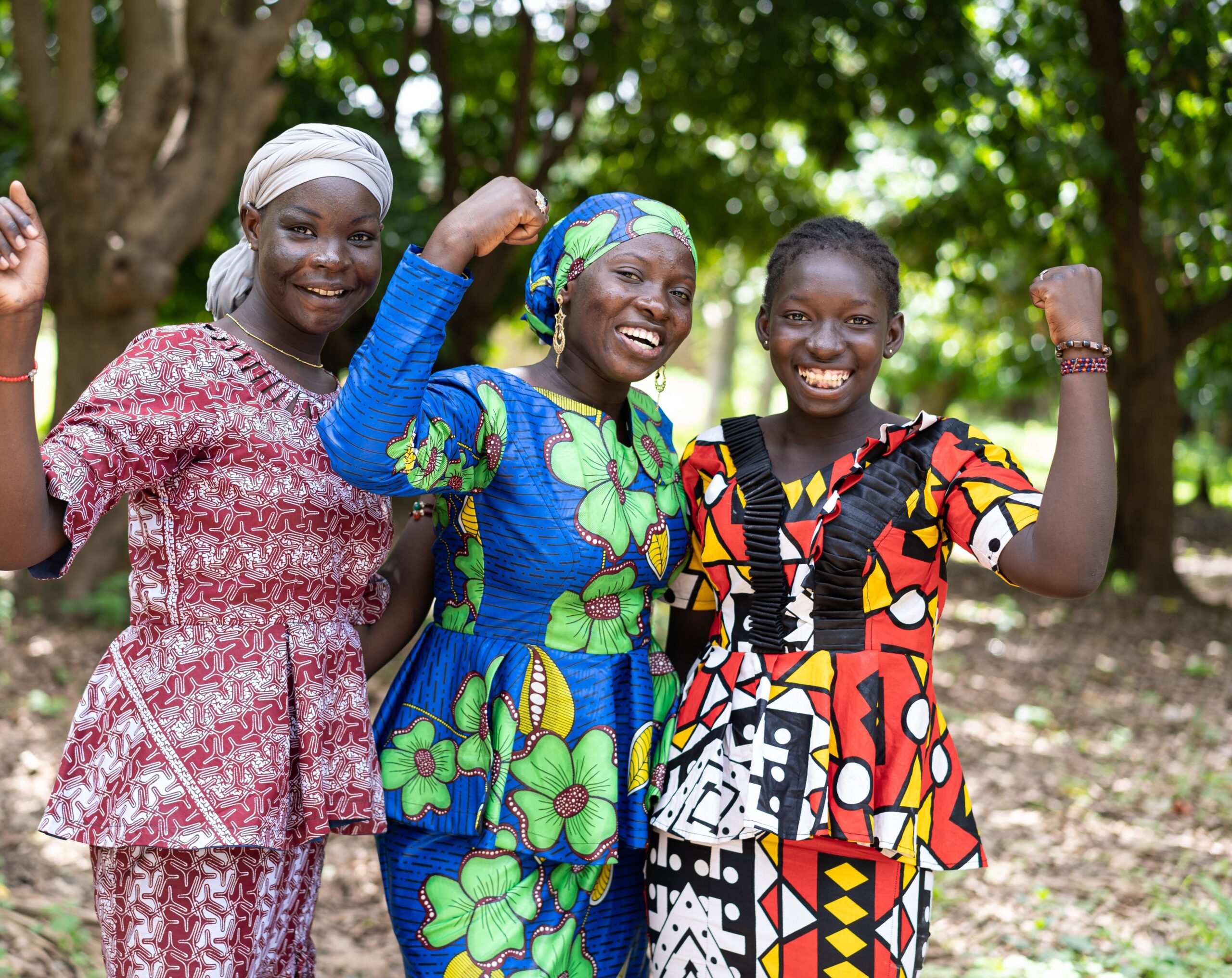 Young African Mother in center with Daughters on either side, all smiling in colorful African print dresses, each with one arm raised in triumph.